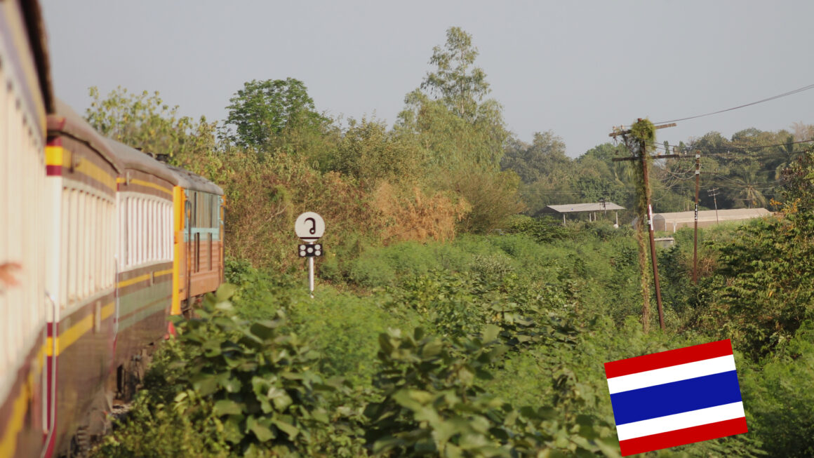 Todeseisenbahn fährt in Rechtskurve durch grüne Landschaft in der Provinz Kanchanaburi. Thailand-Fahne rechts unten im Bild.