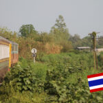 Todeseisenbahn fährt in Rechtskurve durch grüne Landschaft in der Provinz Kanchanaburi. Thailand-Fahne rechts unten im Bild.