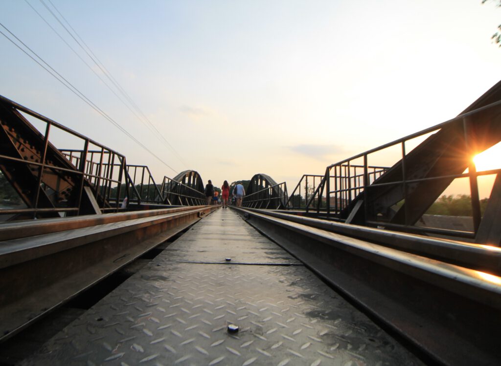 Blick auf Schienenhöhe über die Brücke am Kwai. Im Hintergrund spazieren Touristen auf den Gleisen der Death Railway. 