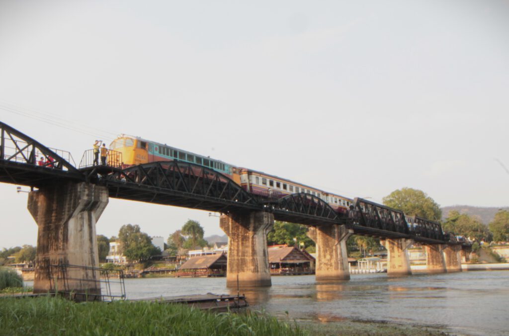 Ein Zug der Todeseisenbahn fährt über die Brücke am Kwai. Schaulustige stehen auf Aussichtsplattformen am Rand der Brücke. Fotografiert vom Kwai-Ufer aus. 