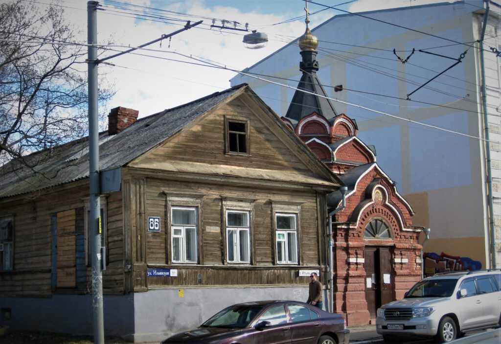 Altes Holzhaus neben einer roten Backstein-Kapelle an einer Hauptstraße in Nischni Nowgorod. 