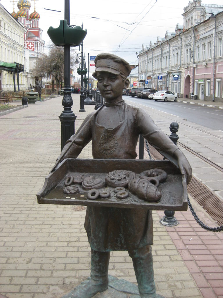 Bronzefigur des Brötchenjungen an Hauptstraße. Junge mit Bäckermütze hält ein Tablett mit verschiedenen Gebäckstücken.
Мальчик с бубликами