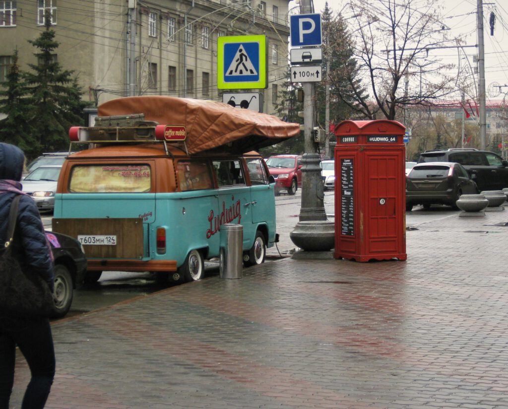 Hellblauer VW-Bus mit brauner Markise an Straßenrand in Krasnojarsk. Aufschrift "Coffee Mobile". Daneben rote Telefonzelle mit Kaffee-Menü. 