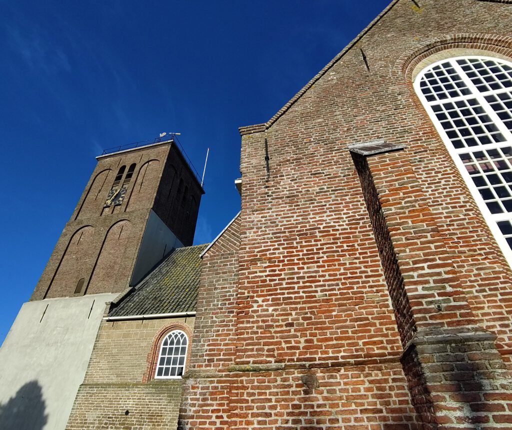 Nahaufnahme der Außenfassade der Maartenskerk in Texel. Rechts Seitenschiff aus rotem Backstein. Links der rechteckige Kirchturm. Dazwischen altes Schiff aus Tuffstein. 