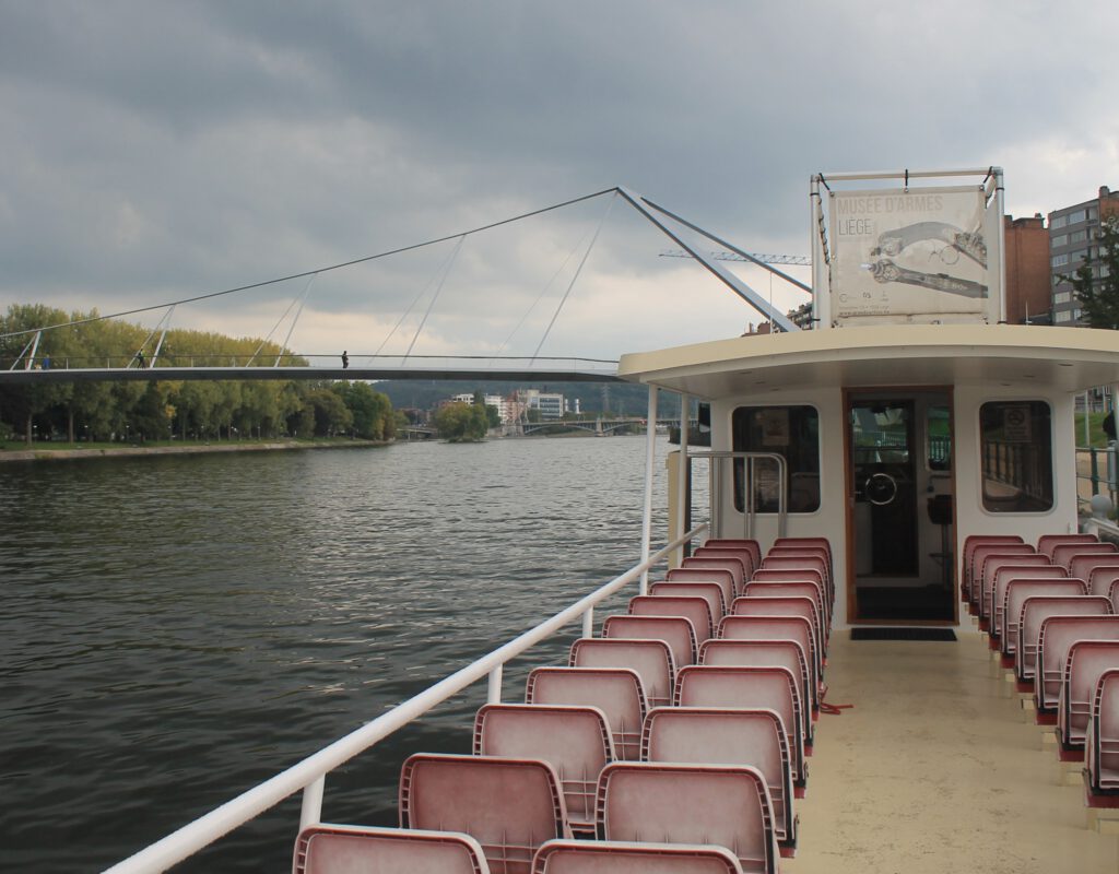 Bootstour auf der Maas. Blick vom Boot aus auf die Maas in Lüttich. Im Hintergrund die Fußgängerbrücke C. Im Vordergrund leere Platiksitze auf dem Außendeck.