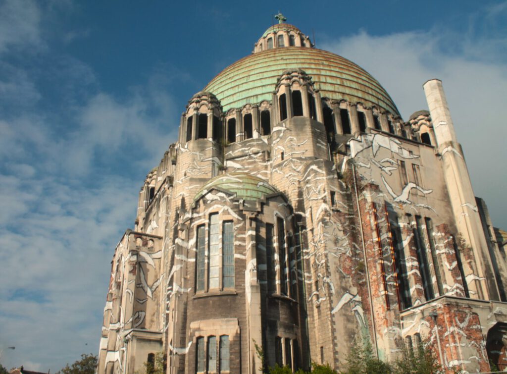 Die Église du Sacré-Coeur de Cointe. Die Kirche ist in Nahaufnahme zu sehen. Ein imposantes Gebäude aus grauem Stein und einer bronzen-grünen Kuppel. Auf der Fassade sind viele weiße Möwen-Graffiti zu sehen. Der Himmel ist blau, mit einigen weißen Wolken.