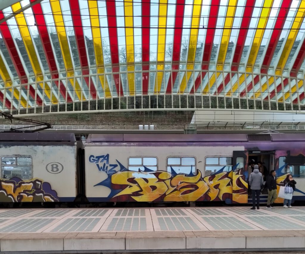 Mit Graffiti bemalter Regionalzug am Bahnhof Liege-Guillemins. Leute steigen ein und aus. Darüber das bunte Dach des Bahnhofs.