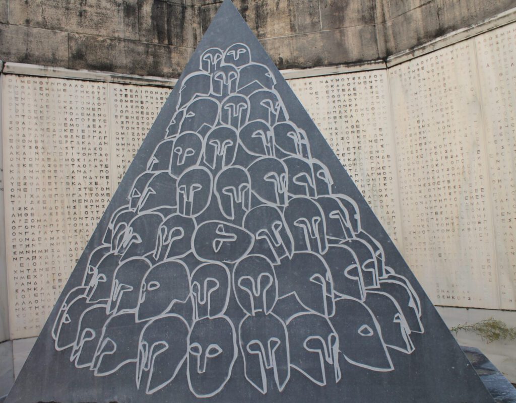 Schwarze Granitpyramide, in die antike Soldatenhelme eingraviert sind. Dahinter eine Wand mit eingravierten alt-griechischen Buchstaben. 