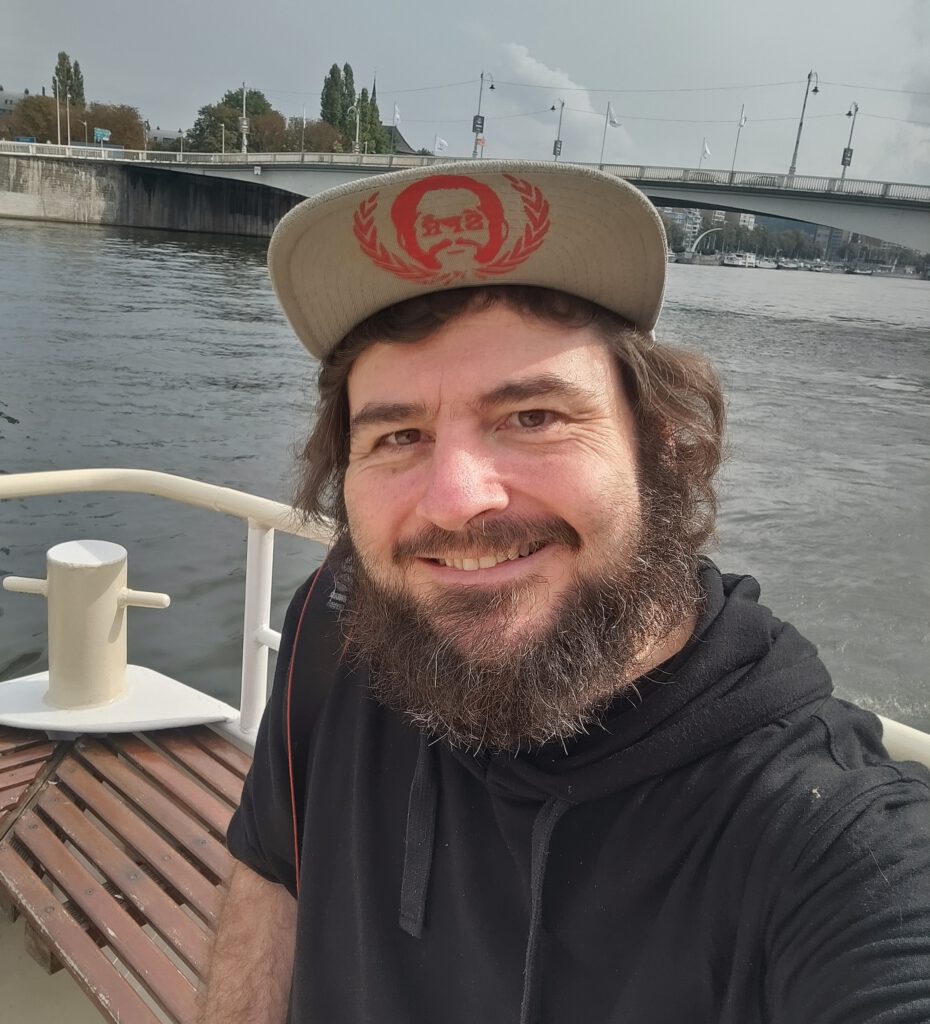 Selfie des Autors auf einem Boot der Navette Fluviale auf der Maas in Lüttich. Im Hintergrund Wasser und eine Brücke.