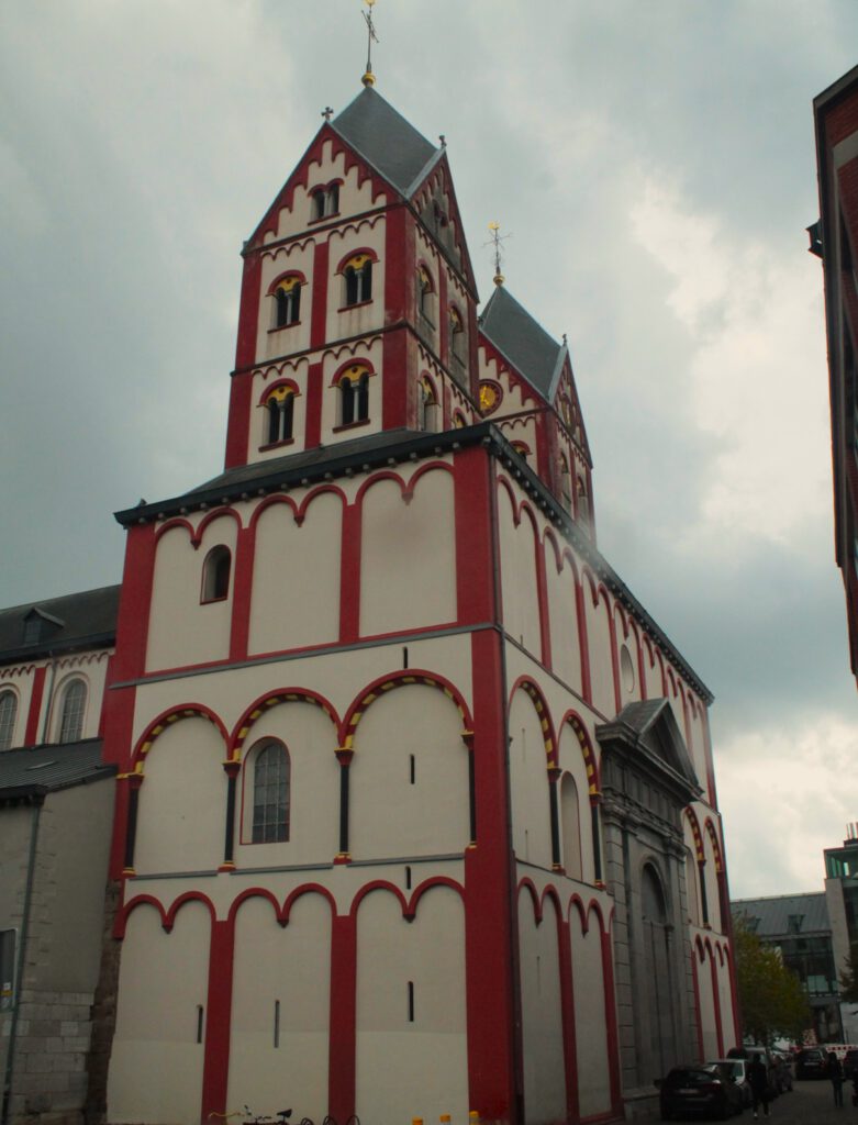 Halb-seitliche Ansicht der Kirche St. Barthelemy in Lüttich. Rot-weiße Fassade mit zwei Türmen, die rhombische Dächer tragen. 