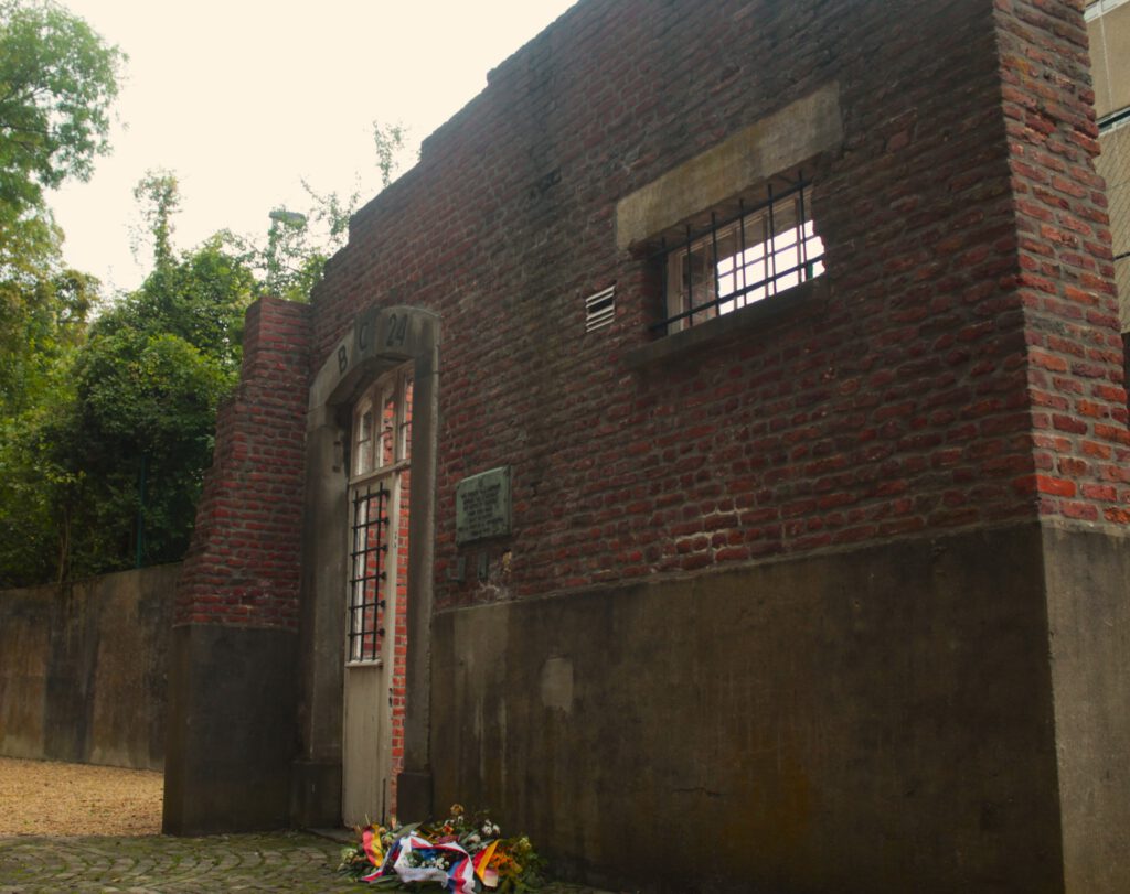 Frei stehende Mauer mit Tür und Fenster auf dem Hof. Davor liegen Gedenksträuße.