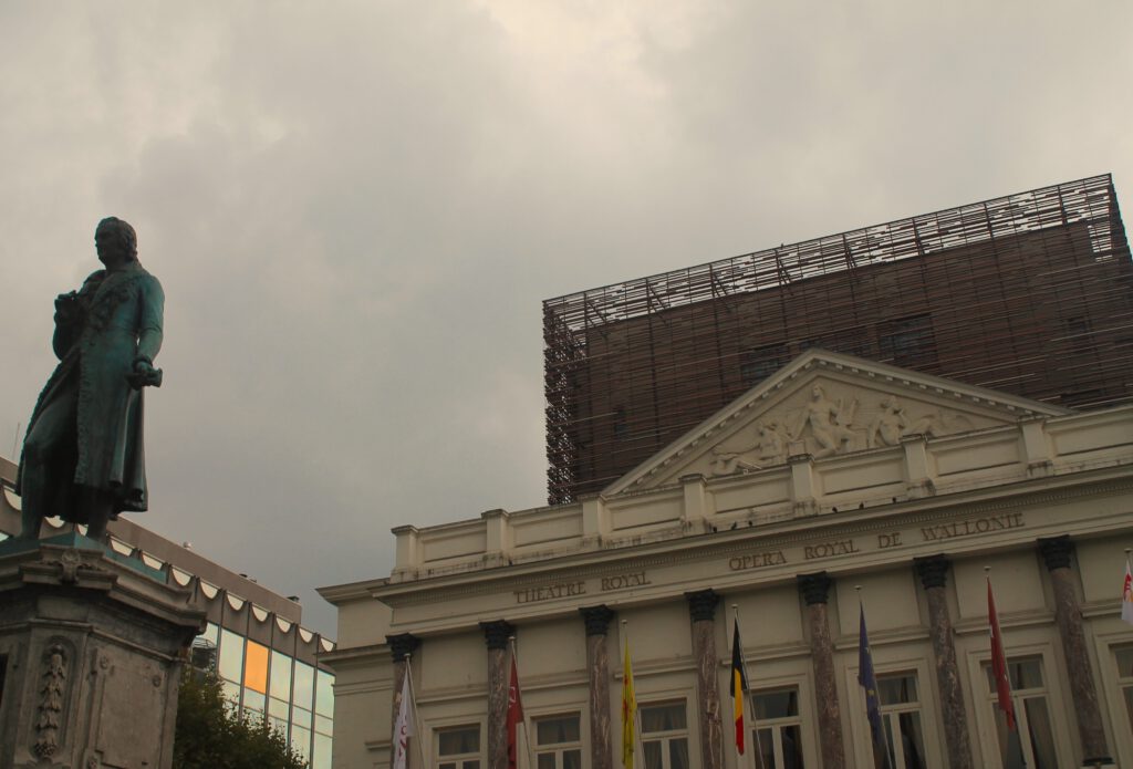 Vorderseite der Opéra Royal de Wallonie. Neoklassizistisches Gebäude mit modernem Anbau, der über die Frontfassade hinausragt. Auf dem Vorplatz eine Bronzestatue auf Betonsockel. 