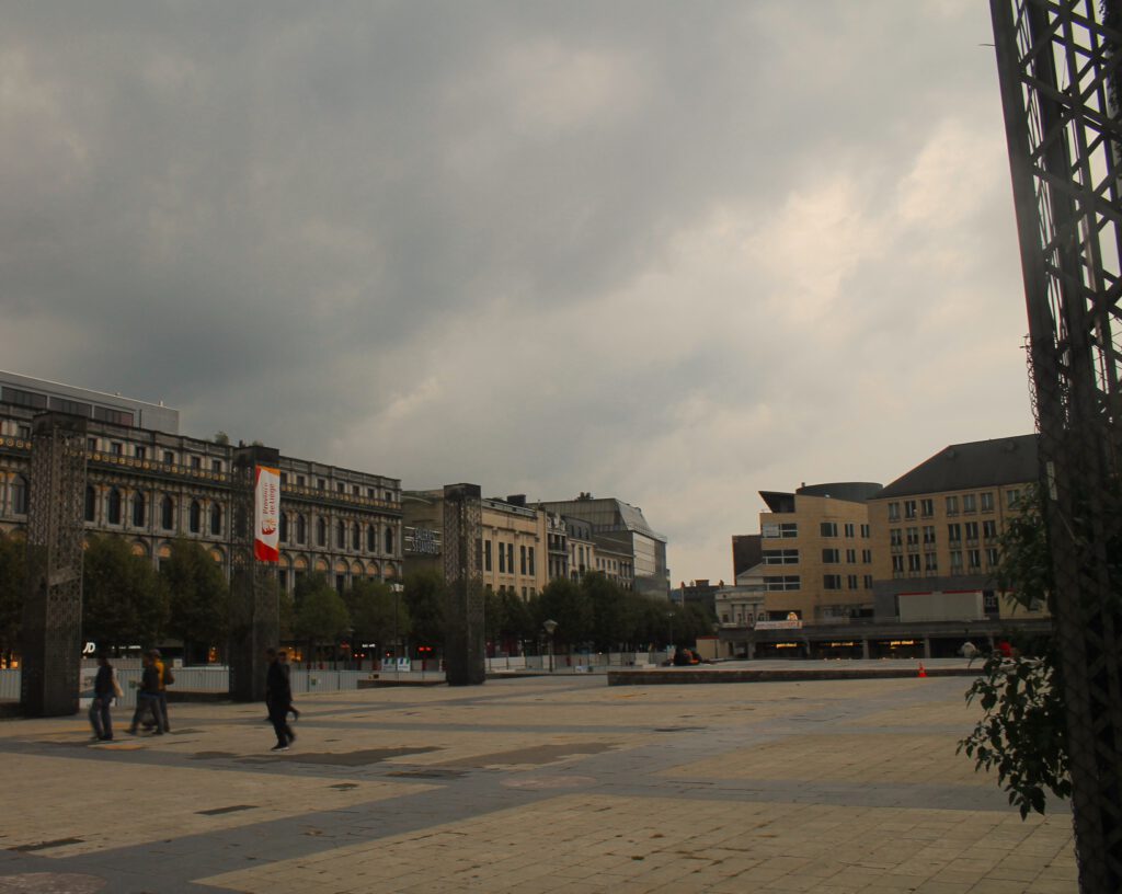 Place Saint Lambert in Lüttich, aufgenommen aus einer ebenen Perspektive. Links im Bild drei der Stahlsäulen. Nur wenige Fußgänger. Grauer Himmel.