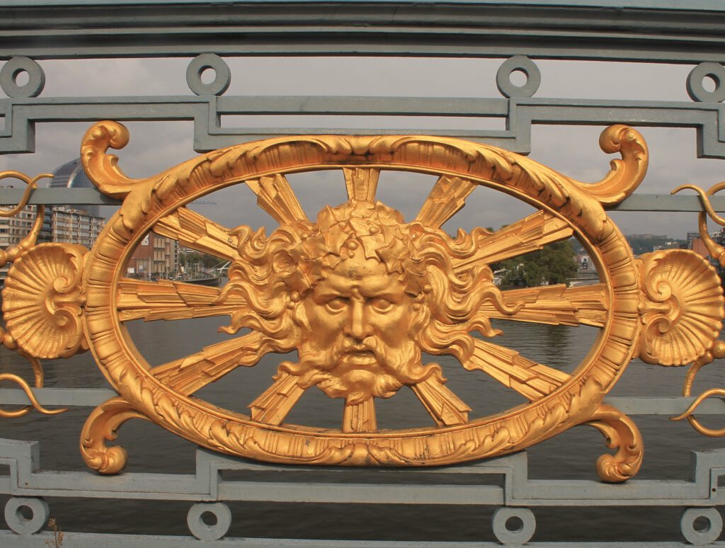 Nahaufnahme einer vergoldeten Bronzeplakette am Geländer der Pont de Fragnee in Lüttich. Sie zeigt das Gesicht einer männlichen, mythogischen Figur mit Schnauzbart. 