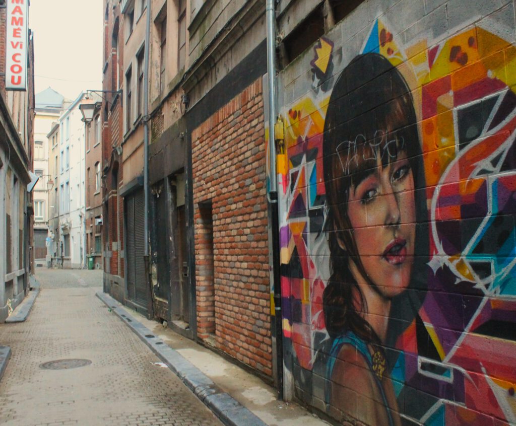 Buntes Graffiti rechts an einer Häuserwand in einer schmalen Gasse in Lüttich. Es zeigt das Gesicht einer jungen Frau vor buntem Hintergrund. 