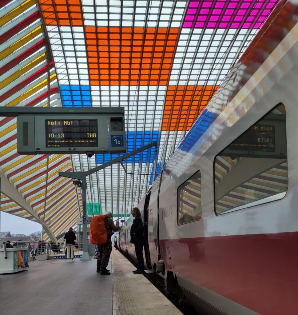 Thalys-Zug auf Gleis 1 am Bahnhof Liege-Guillemenins. Schaffner an der Tür, Gast mit Rucksack steigt zu. Beide seitlich zu sehen. Anzeige: Köln Hbf 10:13