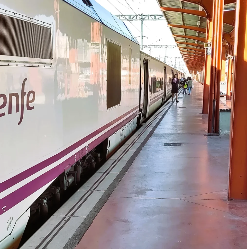 Bahnsteig in Madrid mit weißem Alvia Zug im Gleis. Schaffnerin vor einer Tür. 