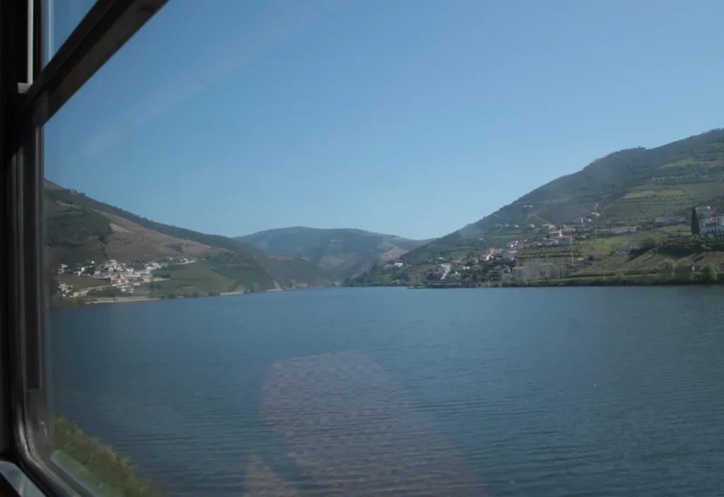 Blick aus Zugfenster auf breiten Douro-Fluss mit hügeligem Ufer.