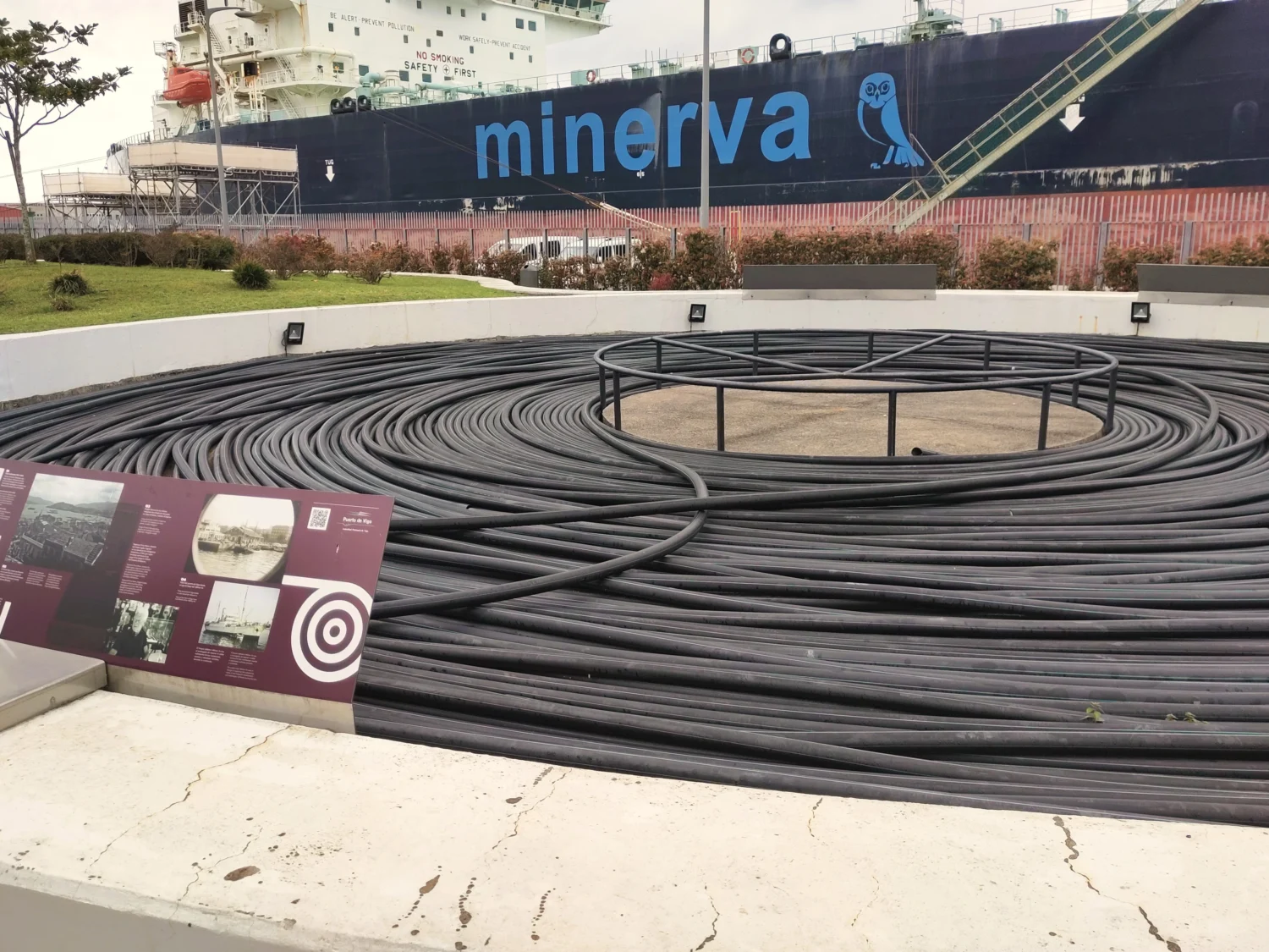 Im Kreis aufgerollte, schwarze Kabel am Hafen von Vigo. Mit Infotafeln. Im Hintergrund ein großes Frachtschiff 
