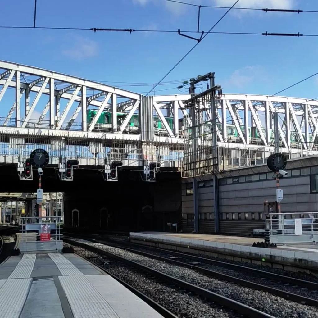 Metallbrücke mit türkisem Metro-Zug über Bahnhofsgleisen.