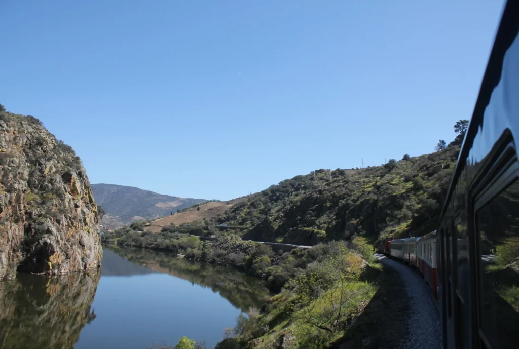 Zug fährt am Douro entlang des Douro Valley. Steinküste und Ufer spiegeln sich im Wasser.